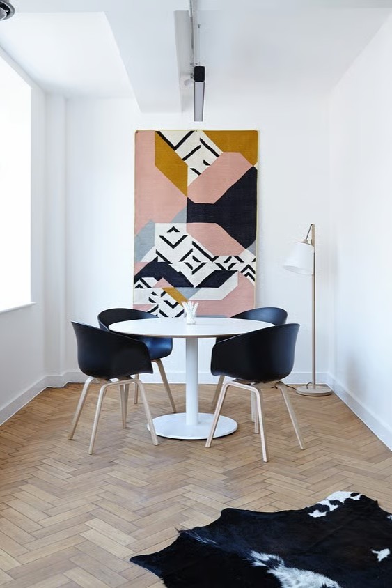 Rincón de reunión moderno, decoración minimalista con paredes blancas, mesa redonda, sillas negras y una alfombra colgante con diseño gráfico abstracto.